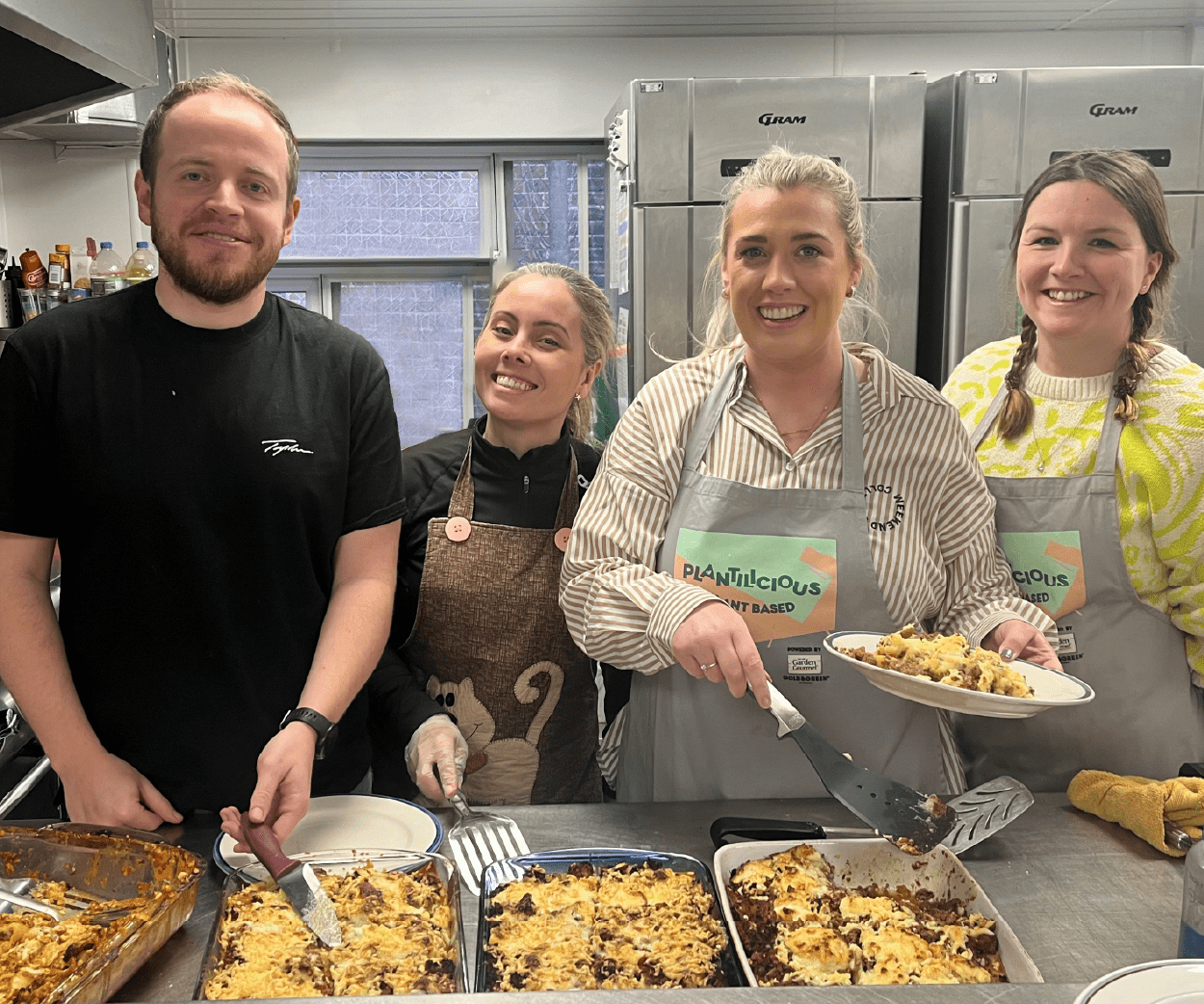 Four smiling people in aprons serving food in a kitchen